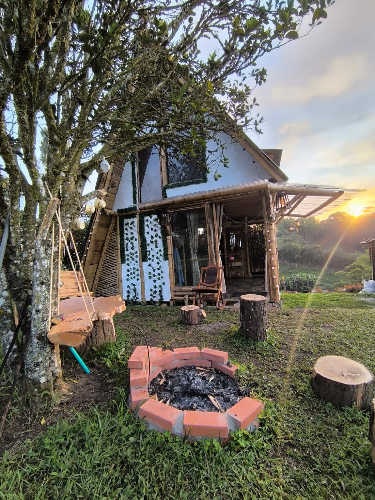 An eco-sustainable cabin is depicted, featuring a unique exterior and warm wooden elements. A fire pit surrounded by a circular brick border is placed on the grassy area, complemented by wooden seating. The setting is highlighted by lush greenery and a sunset sky.