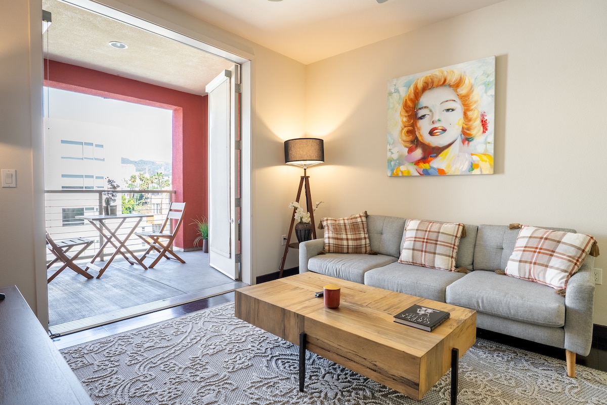 A cozy living room features a light-grey sofa adorned with decorative pillows, a wooden coffee table at the center, and a vibrant wall art piece above. Large glass doors provide access to an outdoor balcony with a small dining set and potted plants.