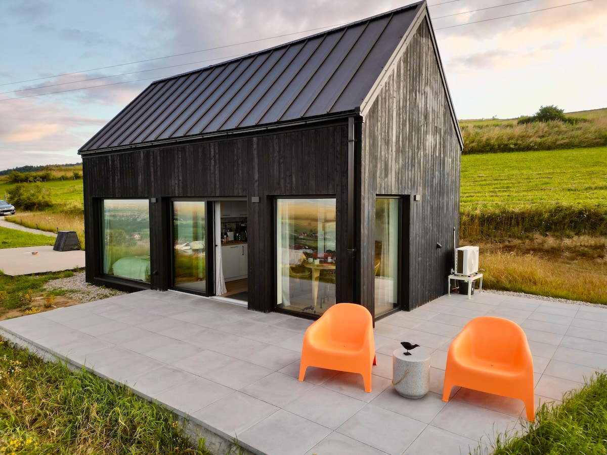 A modern cottage features a sleek black exterior with large glass windows, allowing for natural light. On the outdoor terrace, two vibrant orange chairs are positioned beside a small table, overlooking the green landscape and hills in the background.