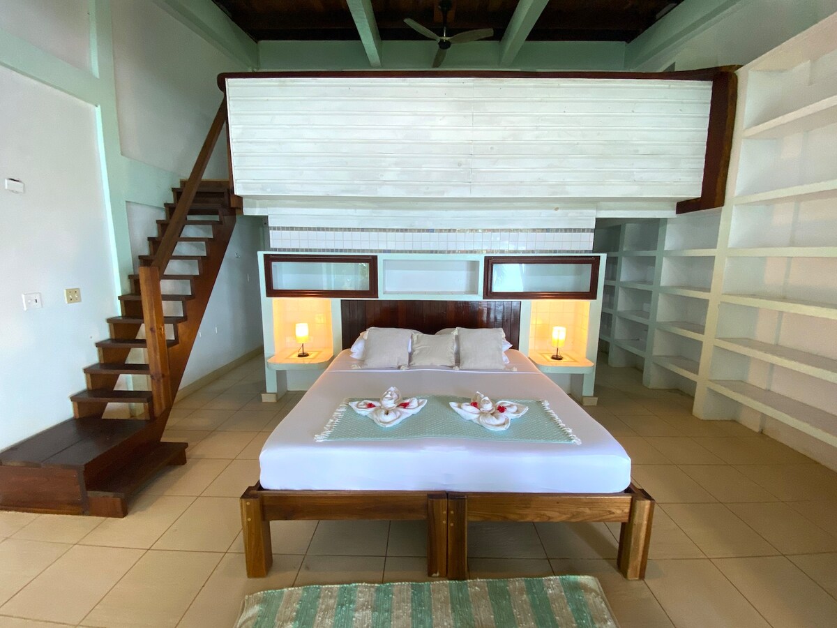A spacious loft suite is presented with a king-sized bed featuring decorative towels arranged on the bedding. A wooden staircase leads to an upper loft area, while built-in shelves and side tables provide additional storage and lighting. Large windows enhance natural light in the room.