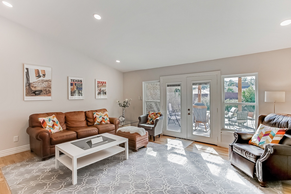 A spacious living area features two brown leather sofas and a light-colored coffee table at its center. Colorful throw pillows add accent to the seating. Natural light floods in through large windows and double doors, which lead to an outdoor seating area.