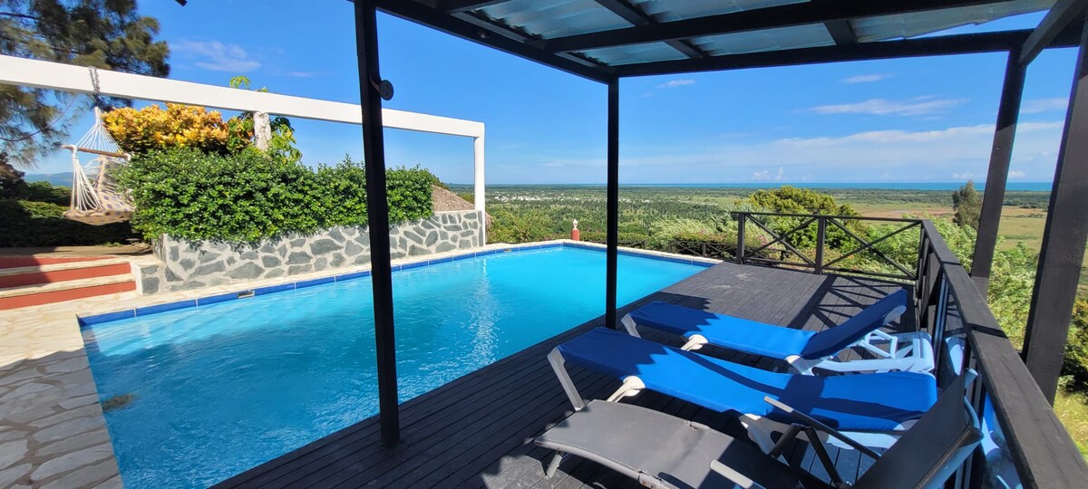 A spacious outdoor pool area features a well-maintained swimming pool with a wooden deck. Comfortable loungers are arranged by the poolside, while lush greenery and a distant view of the coastline enhance the serene atmosphere. A covered structure provides shaded seating nearby.