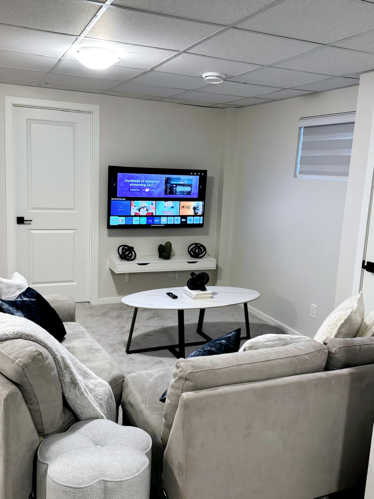 A comfortable living area is presented with a soft, neutral-toned sofa and armchair positioned around a small coffee table. A wall-mounted television displays various streaming options. Light filters in through a window covered by a minimalist blind, enhancing the modern decor.