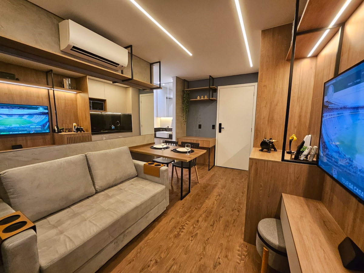 A modern living space is presented with light wood accents and sleek furnishings. A comfortable sofa and a work desk combine functionality with style. The room features a wall-mounted TV, ample lighting, and large mirrors, contributing to an open and inviting atmosphere.