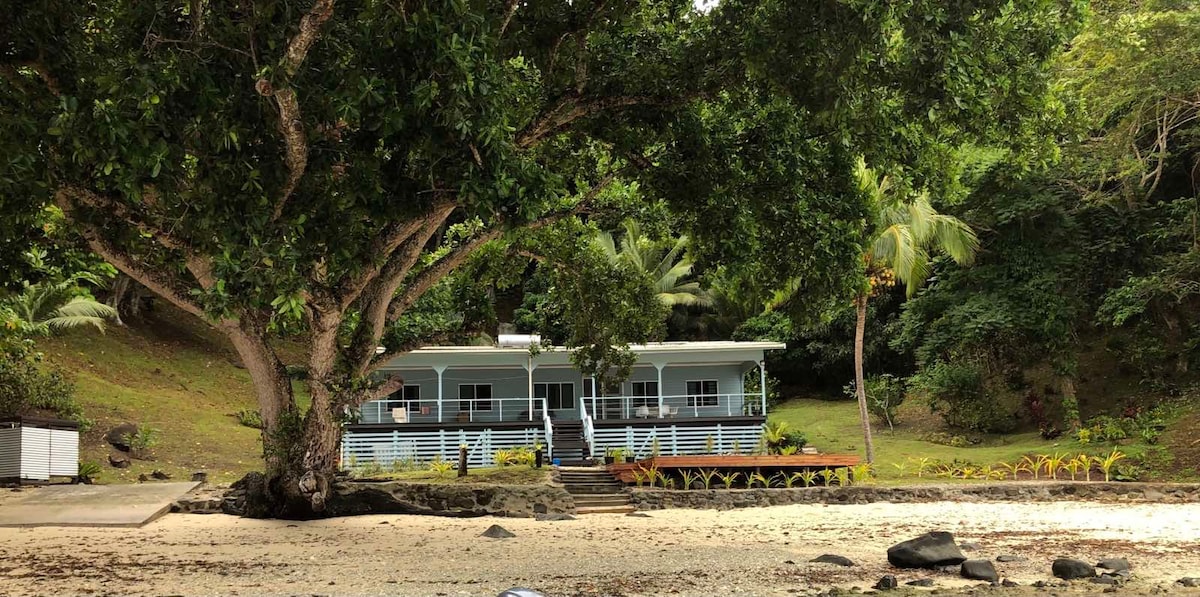 A family-run holiday home is nestled amidst lush greenery, showcasing a welcoming facade with a wide deck. Palm trees surround the property, providing shade and a tropical feel. The sandy beach is visible in the foreground, enhancing the serene coastal ambiance.