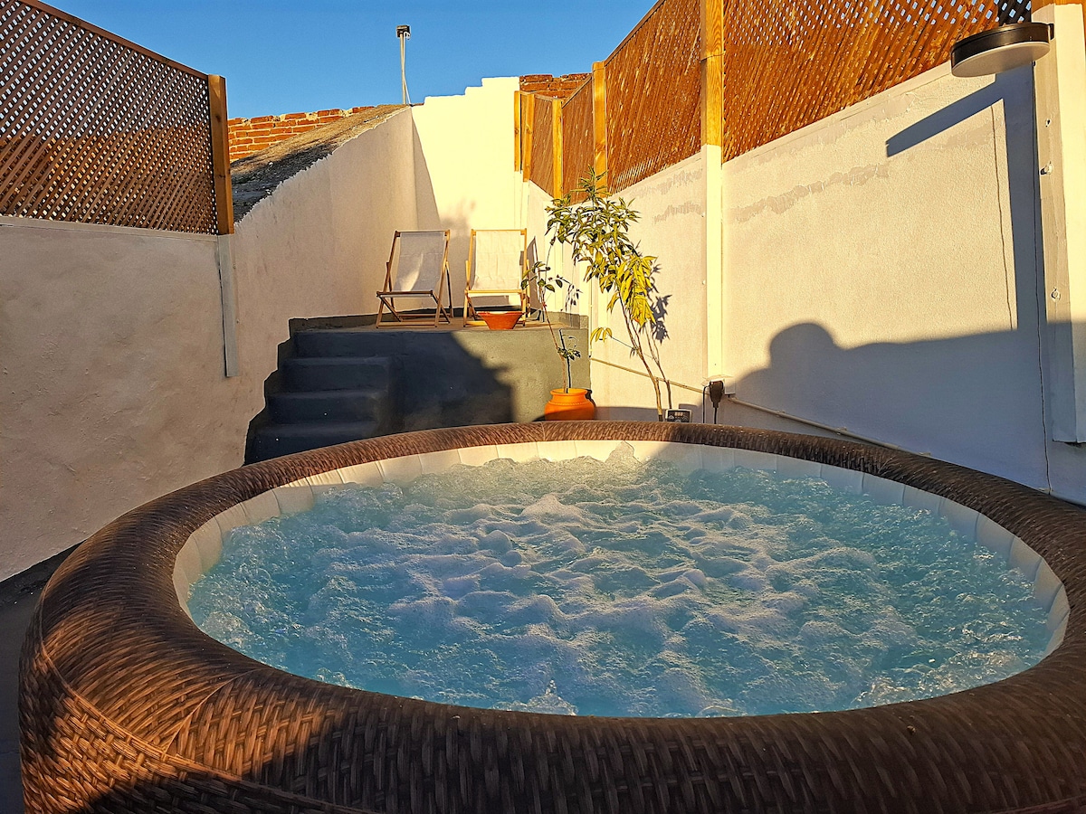 A private outdoor area features a round jacuzzi filled with bubbling water, surrounded by wooden fencing for privacy. Sun loungers are positioned nearby on a set of steps, with a small plant adding a touch of greenery to the space.