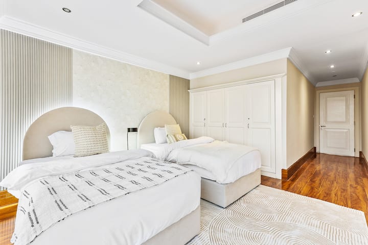 Un dormitorio luminoso con dos camas individuales, cabeceros de color beis, un gran armario blanco y suelo de madera.