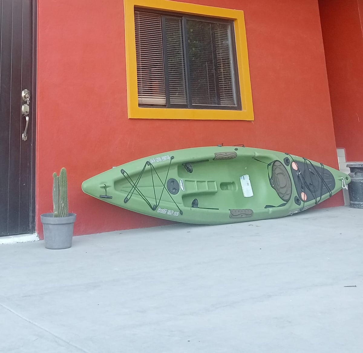 A green kayak is positioned against a vibrant orange wall, accompanied by a potted cactus on the ground. A window with dark framing is visible nearby, contributing to the lively exterior setting.