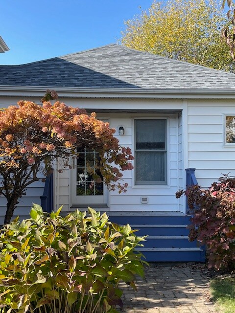 The exterior features a charming white house with a sloped gray roof and blue steps leading to the entrance. A vibrant hydrangea bush with autumn foliage is showcased in the foreground, while an inviting tree complements the scene.