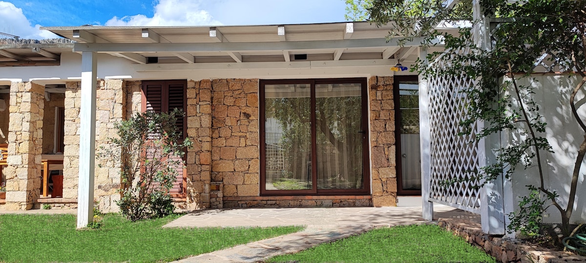 The exterior of the apartment is featured, showcasing a charming stone facade and large glass windows. Lush green grass surrounds the entryway, and a covered porch is visible, providing shaded outdoor space. Nearby foliage adds to the home’s tranquil setting.