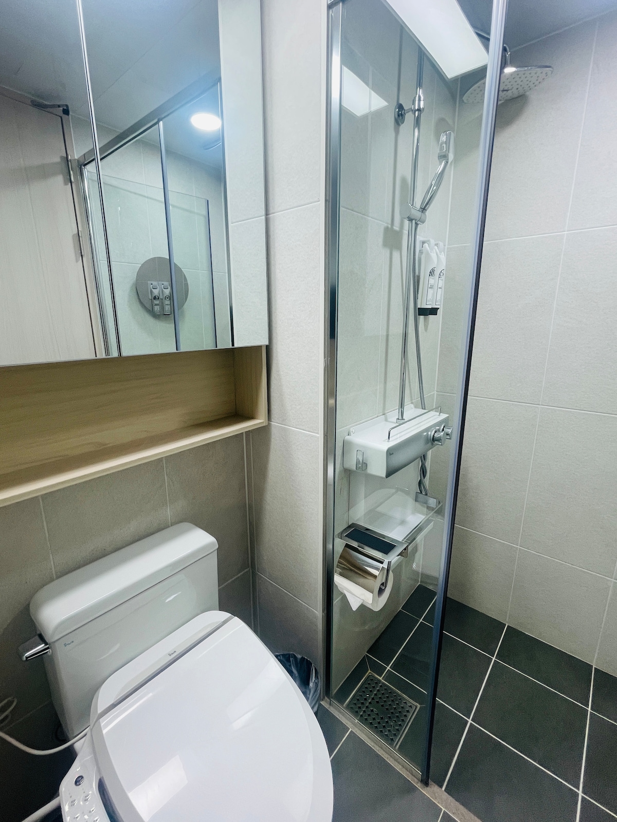 A modern bathroom is presented with sleek tiled walls and a glass shower enclosure. A clean toilet sits beside the shower, and a minimalistic sink with neatly arranged toiletries is visible. Ample lighting reflects off the mirrors, enhancing the room's spacious feel.