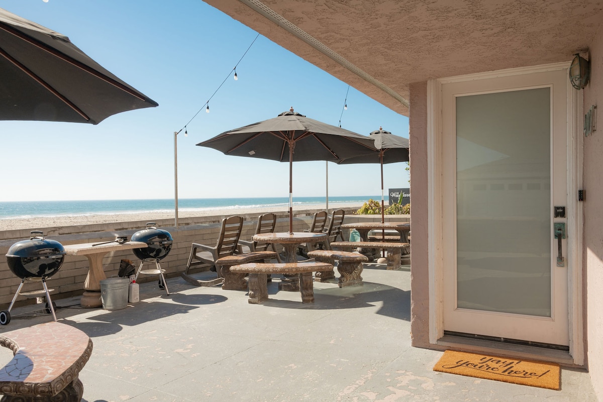 A spacious beachfront patio showcases a large dining table surrounded by natural stone seating and shaded by umbrellas. Two charcoal grills are positioned nearby, with direct views of the ocean sparkling in the sunlight. A welcoming mat lies at the entrance, inviting guests indoors.