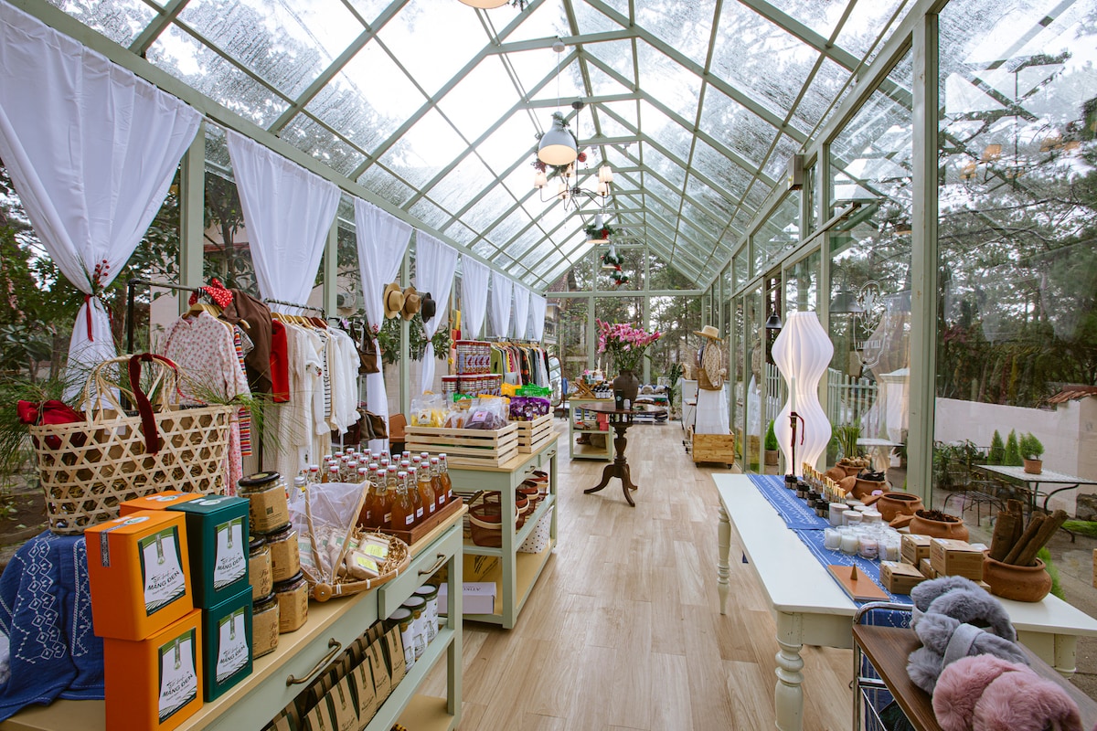 A bright and airy shop is enclosed in glass, showcasing a variety of handmade goods and souvenirs. Shelves display colorful bags, clothing, and local crafts, while light filters through the transparent ceiling. Natural wood floors complement the warm, inviting atmosphere.