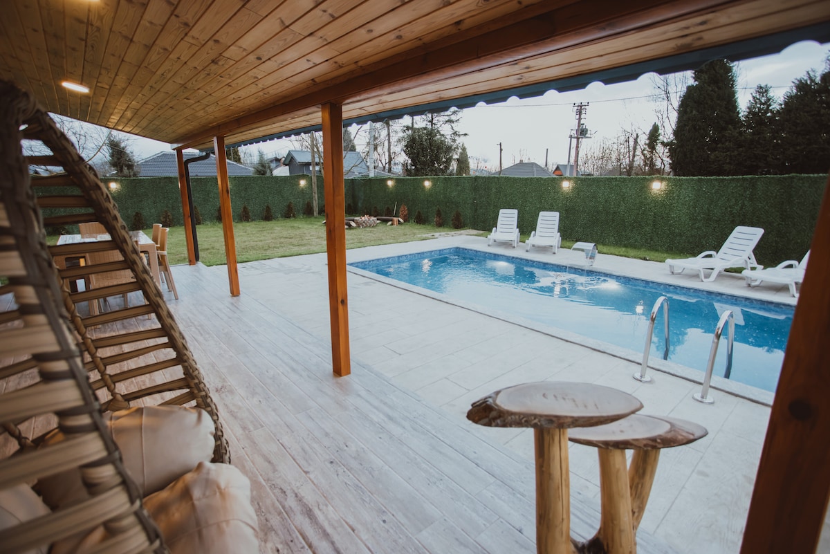 An outdoor area features a spacious pool surrounded by a manicured lawn. Sun loungers are arranged nearby, and a wooden deck covers the floor. Rustic stools and a hanging chair add to the inviting atmosphere, with greenery visible in the background.