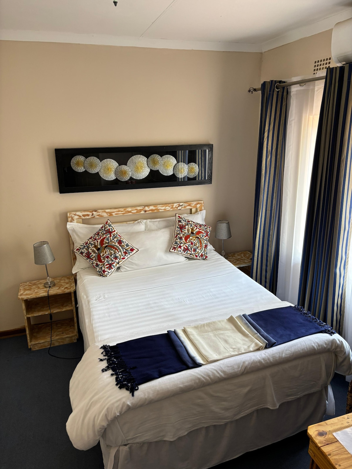 A well-furnished bedroom features a spacious bed with decorative pillows and a light blanket draped at the foot. Two bedside lamps provide soft illumination. A floral wall art piece is displayed above the bed, with window curtains in a striped pattern allowing natural light to enter.
