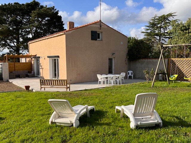 A renovated house is depicted, featuring a patio with outdoor furniture and a grassy yard. Two lounge chairs are positioned on the lawn, offering a relaxed setting. The building's facade is simple, with windows and a door that opens to the outdoors. Trees provide gentle shade.