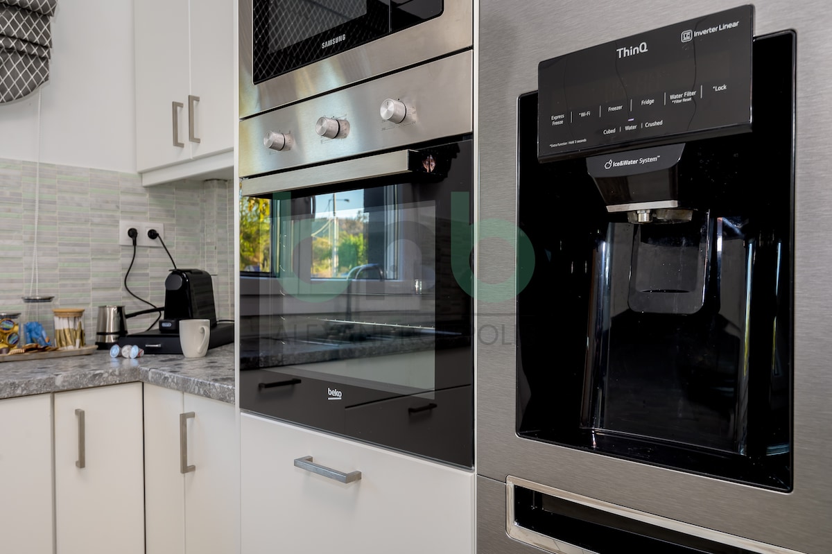 A modern kitchen is showcased with stainless steel appliances, including an oven and refrigerator. The cabinetry features a light finish, while a smooth countertop provides space for a coffee maker and accessories. Natural light is seen entering through a nearby window.