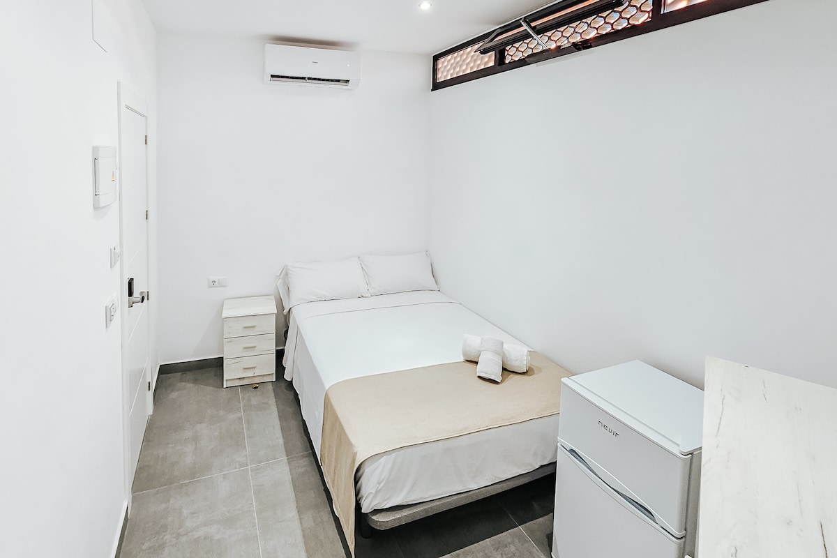 A modern and cozy bedroom is displayed, featuring a spacious bed with neutral bedding. A light-coloured blanket is draped across the bed. A mini fridge and a small bedside table are visible, along with a window that allows natural light to enter the room.