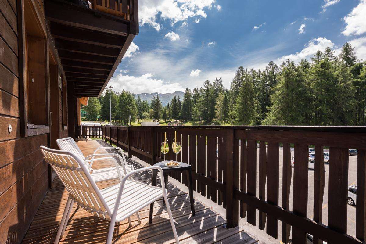 A wooden balcony features two white lounge chairs positioned next to a small table with two glasses and snacks. In the background, trees line the area, and a view of mountains can be seen under a partly cloudy sky.