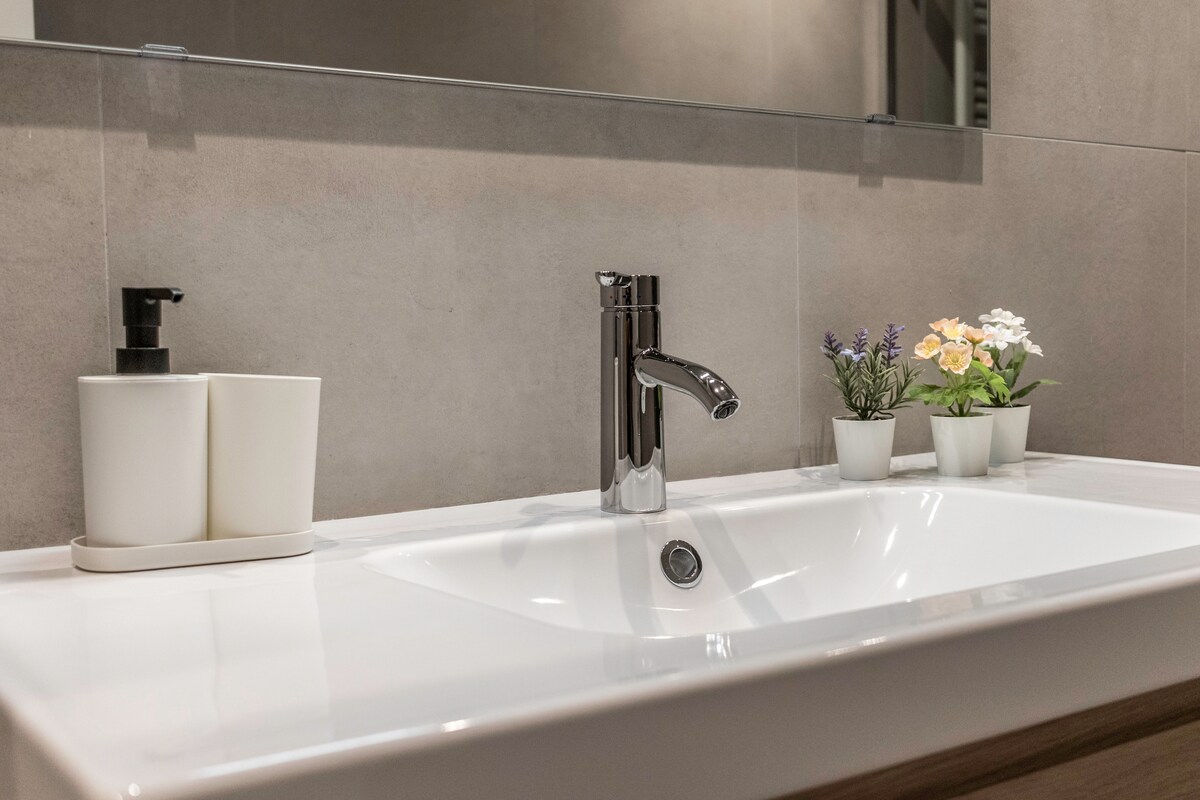 A modern bathroom sink features a sleek chrome faucet, complemented by two decorative plant pots containing small flowers. Nearby, a soap dispenser and a lotion bottle are organized neatly, enhancing the clean and functional aesthetic of the space.