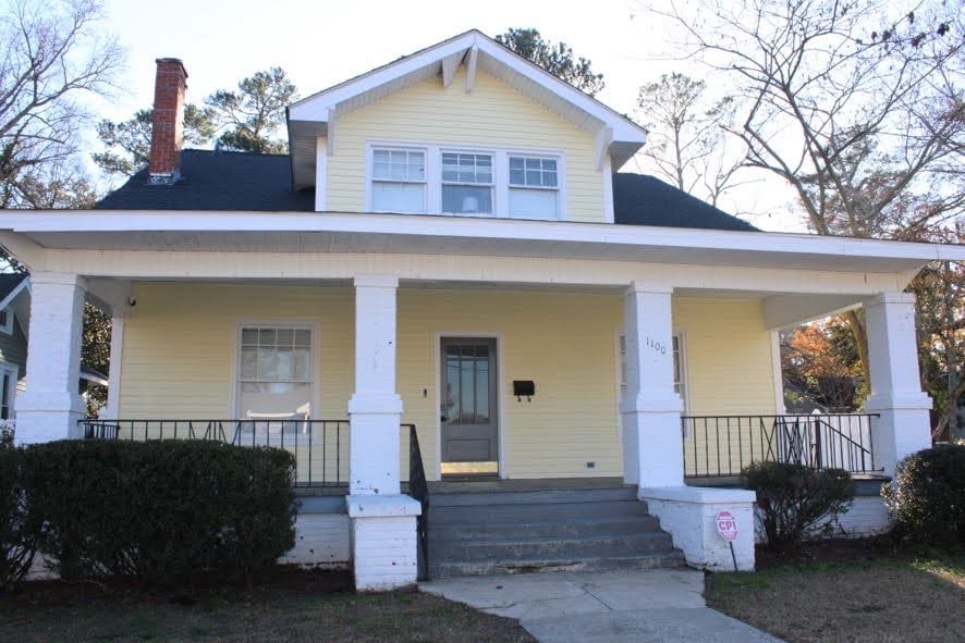 A charming yellow house is displayed, featuring a spacious porch supported by white columns. The front stairs lead to a welcoming entrance, flanked by neatly trimmed bushes. Large windows allow natural light to enter, enhancing the home's inviting façade.
