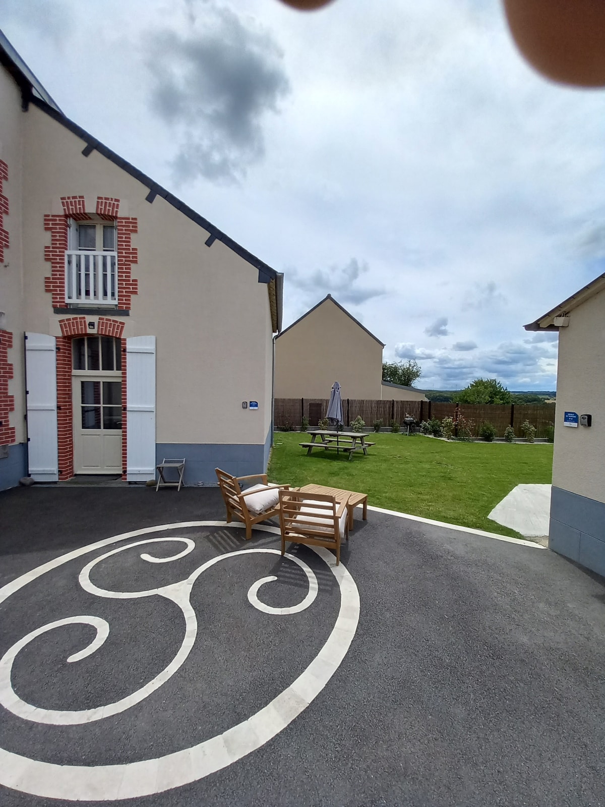 A charming outdoor area features a well-maintained lawn with a picnic table and an umbrella for shade. Two wooden chairs are positioned on a decorative pavement outside the entrance, offering a relaxed seating option. A gentle view of the surrounding countryside is visible in the background.