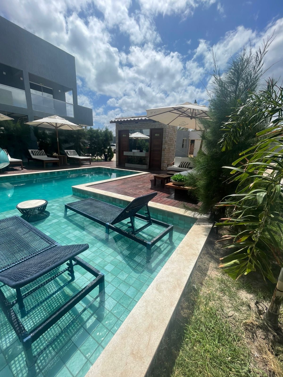 A sunlit pool area features two lounge chairs positioned beside a clear turquoise pool. A shaded seating area with umbrellas and a barbecue space is visible in the background, surrounded by lush greenery and a well-maintained lawn.
