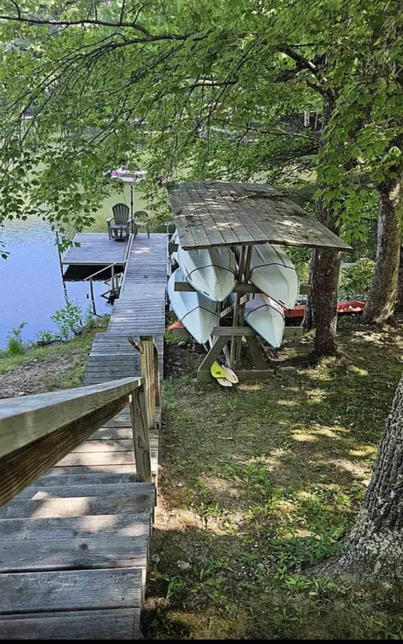 A serene lakeside scene features a wooden dock leading to calm waters, flanked by leafy trees. A storage rack displaying several kayaks stands beside the dock, with an empty chair poised nearby for relaxation. Natural verdant surroundings enhance the peaceful atmosphere.