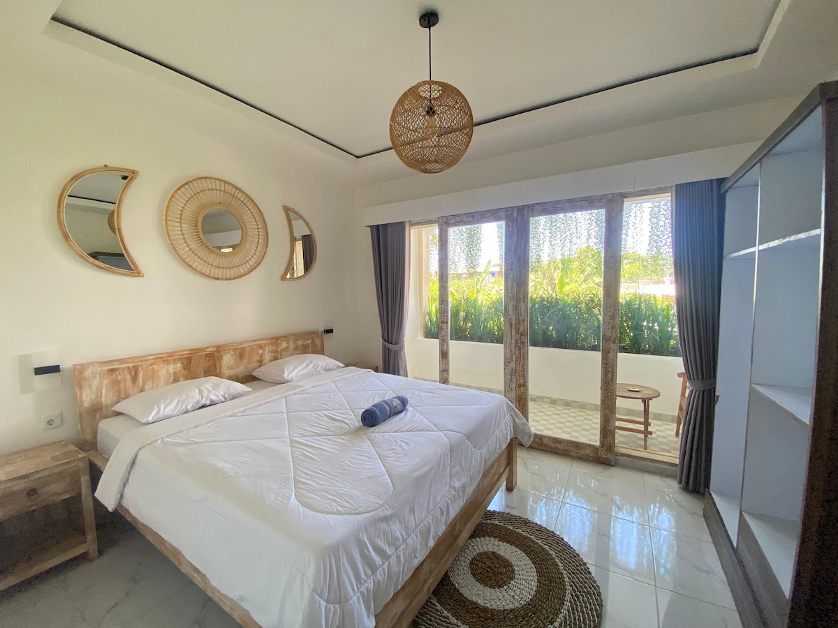 A modern bedroom features a spacious layout with two beds, styled with neutral bedding and decorative towels. Natural light floods through large glass doors that open to a green view. Round mirrors and a woven light fixture add visual interest to the serene space.