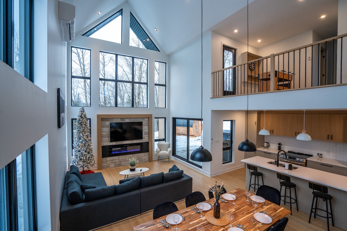 A spacious living area features large windows that allow natural light to fill the room, highlighting a comfortable sectional sofa and a modern dining table set for a meal. A Christmas tree stands elegantly in one corner, while a sleek kitchen is visible in the background.