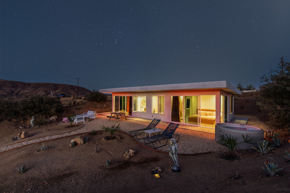 A modern house is illuminated against a starlit sky, showcasing expansive glass windows that offer views of the surrounding desert landscape. Seating areas are visible outside, along with a hot tub positioned next to the structure. The natural terrain features desert plants and rocks.