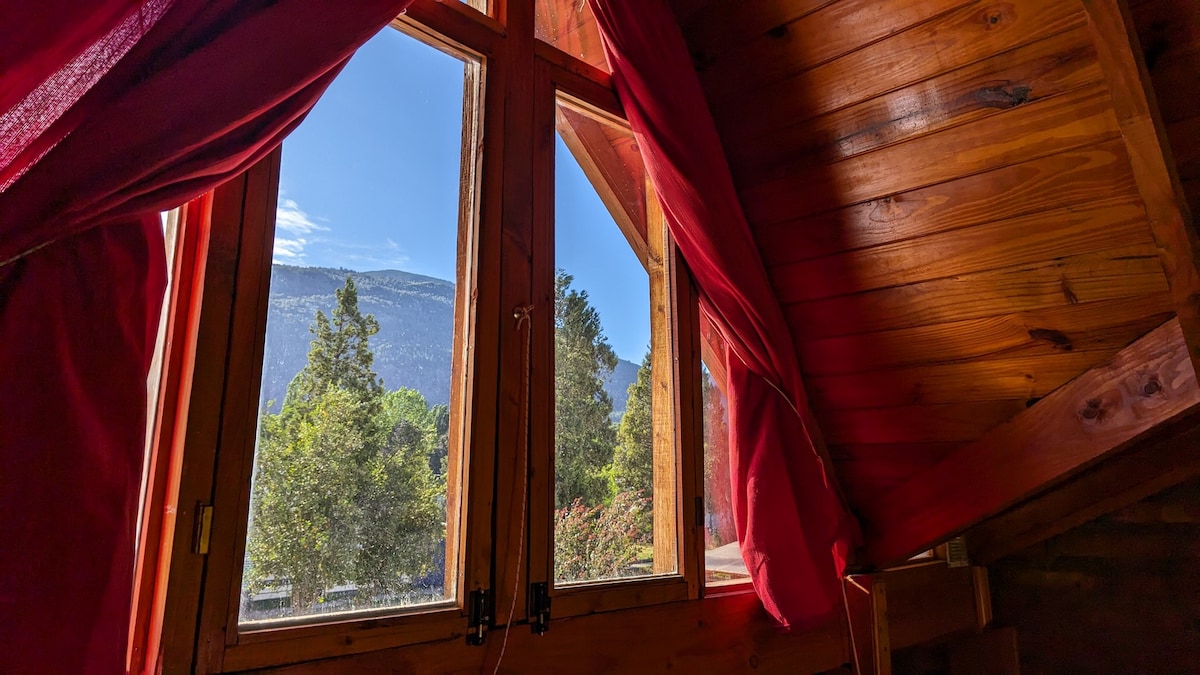 Large wooden windows are framed with rich red curtains, offering a view of the surrounding landscape. The natural light illuminates the wooden interior, enhancing the cabin's cozy design. Lush trees and distant mountains can be seen outside, adding to the serene atmosphere.