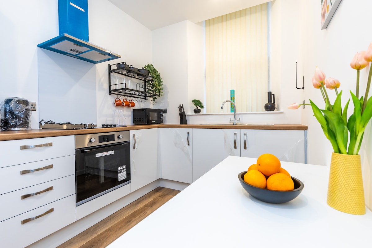 A modern kitchen features sleek white cabinetry and a wooden countertop. An oven and gas hob are included, along with open shelving displaying kitchen essentials. A bowl of oranges is placed on the dining table, while tulips are arranged in a yellow vase by the window.