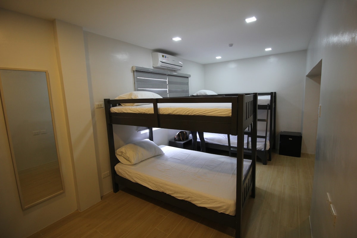 A spacious sleeping area features two sets of sturdy bunk beds, each with simple white bedding. A small black cabinet is positioned nearby, and natural light filters in through a window covered by sleek blinds. A full-length mirror reflects the clean, modern décor.