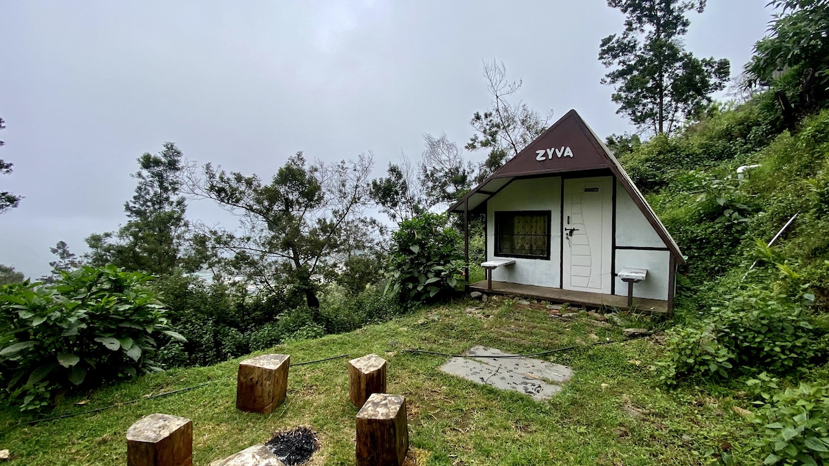 A wooden cabin in a serene setting is framed by lush greenery. A small outdoor seating area with round wooden stools is positioned on a stone surface in front of the cabin. The cloudy sky adds a tranquil ambiance to the scene.