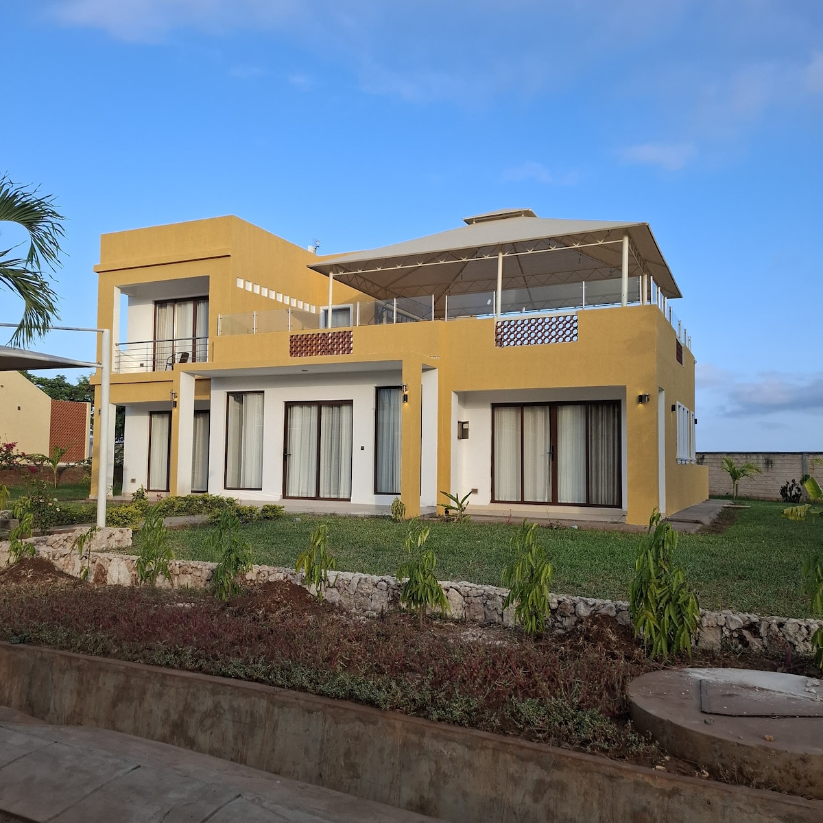 A two-story house features a modern design with large glass doors and a spacious upper terrace. The building is surrounded by well-maintained green grass and landscaping, offering a serene outdoor environment. The exterior walls are painted in a soft yellow hue, blending well with the sky.