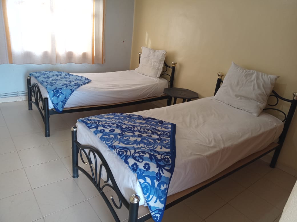 The room features two single beds with decorative blue blankets draped over the corners. A small round table is positioned between the beds. Natural light enters through a window with light, sheer curtains, illuminating the neutral-toned walls and tiled floor.