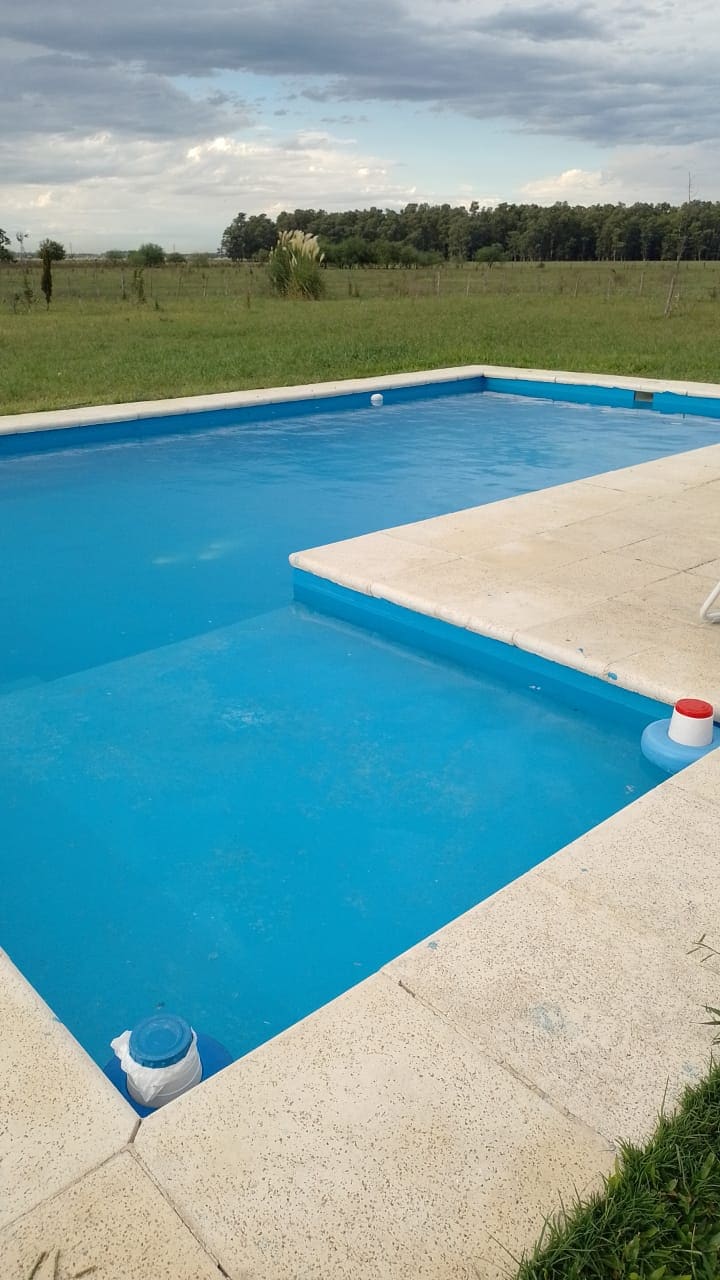 A clear blue pool is surrounded by a spacious concrete deck. The pool area offers views of the lush green field beyond, while nearby accessories add functionality. The sky above features scattered clouds, enhancing the outdoor ambiance.