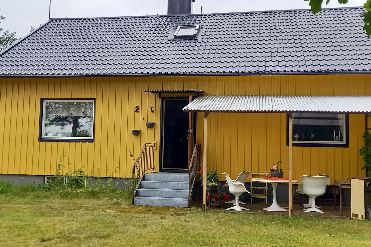 A yellow house with a sloped roof is shown, featuring a covered porch area that includes outdoor seating. A set of stairs leads up to the main entrance, framed by greenery and a well-maintained lawn.