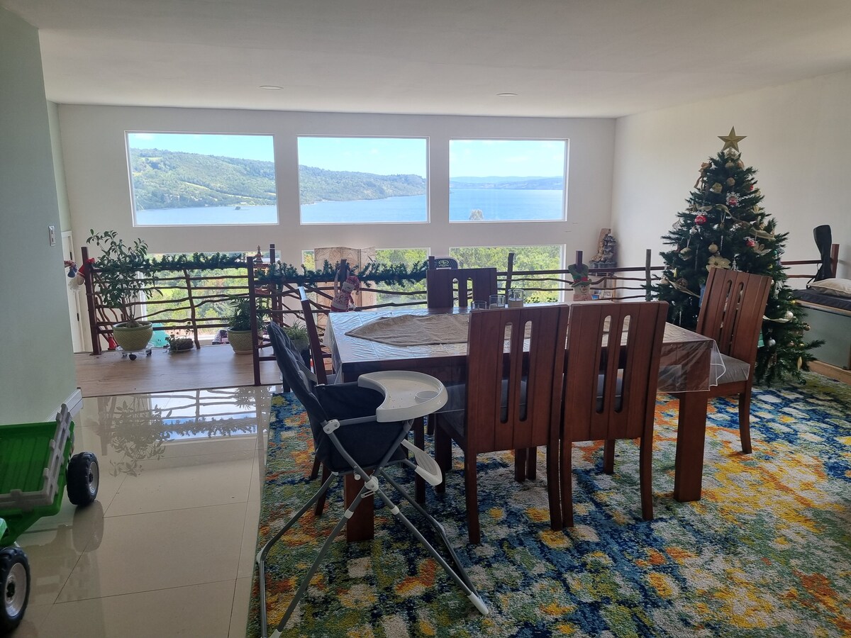 A spacious dining area features a large wooden table surrounded by multiple wooden chairs. Floor-to-ceiling windows offer panoramic views of the lake and surrounding greenery. A decorative Christmas tree stands in one corner, and vibrant rugs add color to the tiled floor.