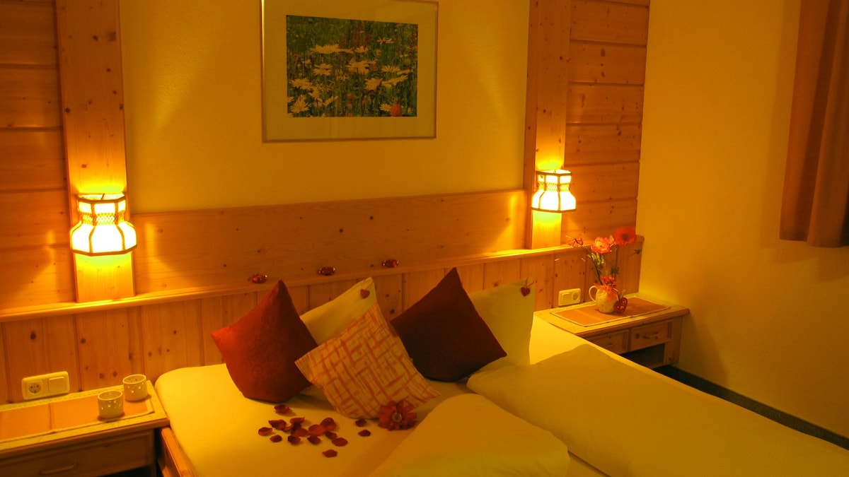 A cozy sleeping area is presented with warm wood paneling and soft lighting. Two spacious beds are adorned with contrasting cushions and decorative elements. The wall features a framed art piece, while bedside tables complement the tranquil ambiance.