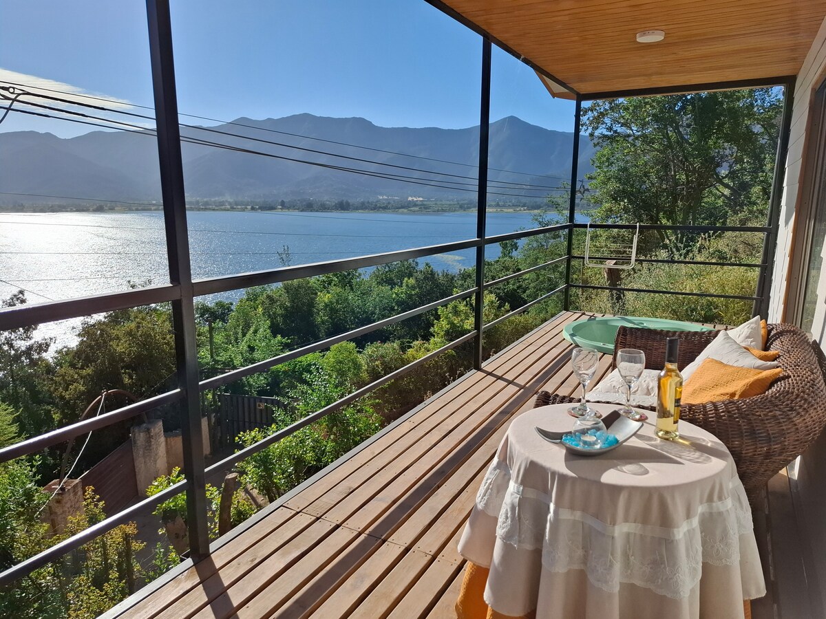 A private terrace offers a serene view of the Aculeo lagoon, framed by distant mountains. A small table is set with two glasses and a bottle, inviting relaxation. Lush greenery surrounds the area, enhancing the connection with nature and the tranquil atmosphere.