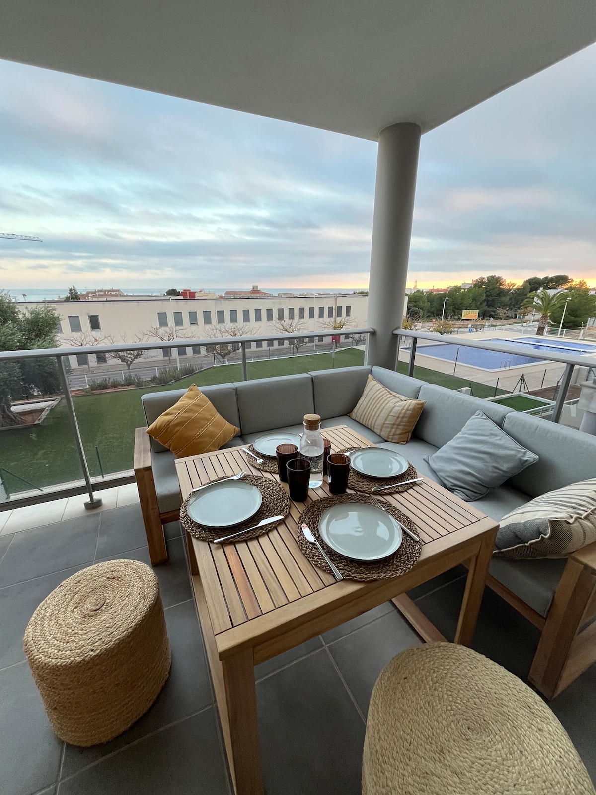 An outdoor dining area features a wooden table set with plates and glasses. Cushioned seating surrounds the table, offering comfort. Natural light fills the space, with a view of nearby greenery and buildings visible in the background.