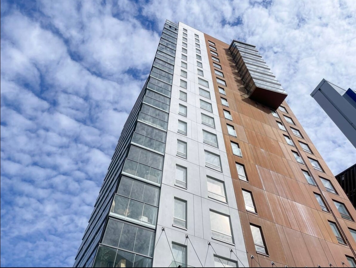 A modern seven-story building is depicted, featuring a blend of glass and textured surfaces. The façade showcases a mix of light and dark elements, with large windows reflecting the sky. A balcony extends from one of the upper-level units, under a backdrop of fluffy clouds.