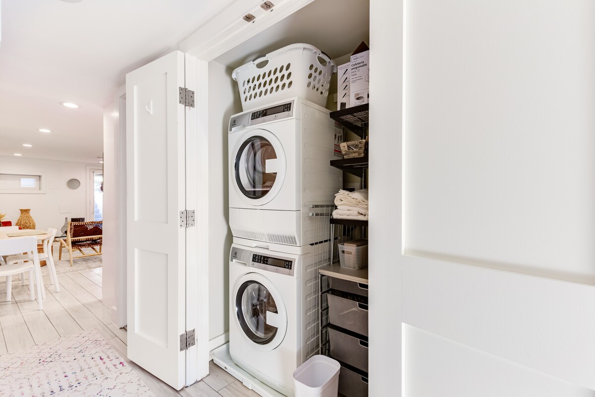 A modern laundry area is featured within a light, compact space. The stacked washer and dryer are neatly arranged beside a basket of laundry. Shelving is filled with additional supplies, while a wastebasket is positioned on the floor for convenience.