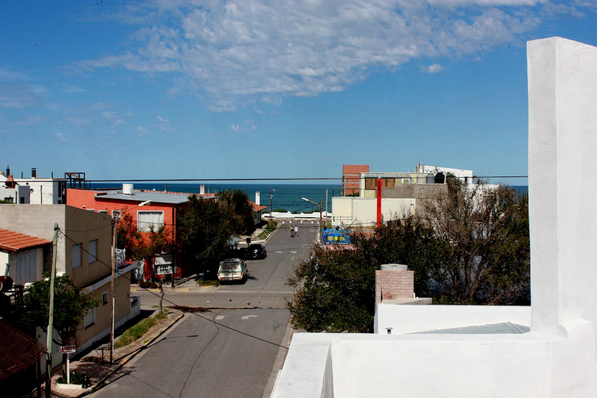 A sea view is presented from a terrace, showcasing the blue waters in the distance and a quiet street lined with buildings. Trees partially obscure the view, while the sky features scattered clouds, contributing to a serene ambiance.
