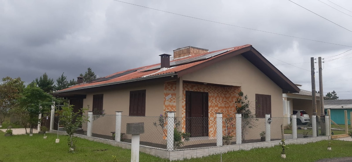 A single-story house is shown with a combination of light and textured brick exteriors. Windows with wooden shutters are visible, while a well-maintained grassy yard features several young trees. A fence surrounds the property, contributing to a sense of privacy.