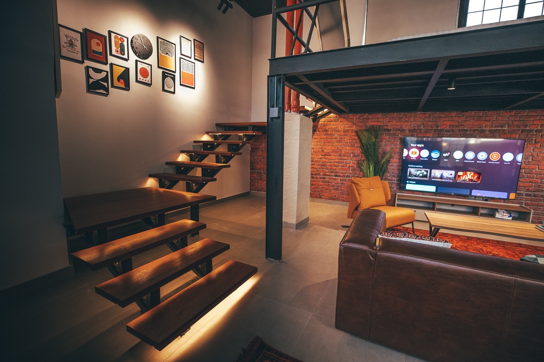 A modern living space showcases a mix of industrial and contemporary design. Wooden stairs lead to an upper level, illuminated by warm lighting. A wall adorned with framed art complements a comfortable seating area that faces a large flat-screen television.