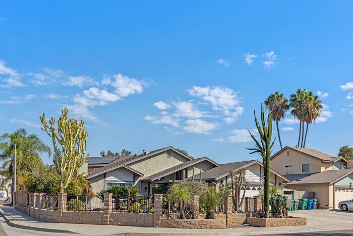 A corner view of a single-family home surrounded by a low wall. The property features diverse desert plants, including tall cacti and palm trees, under a clear blue sky scattered with a few clouds.