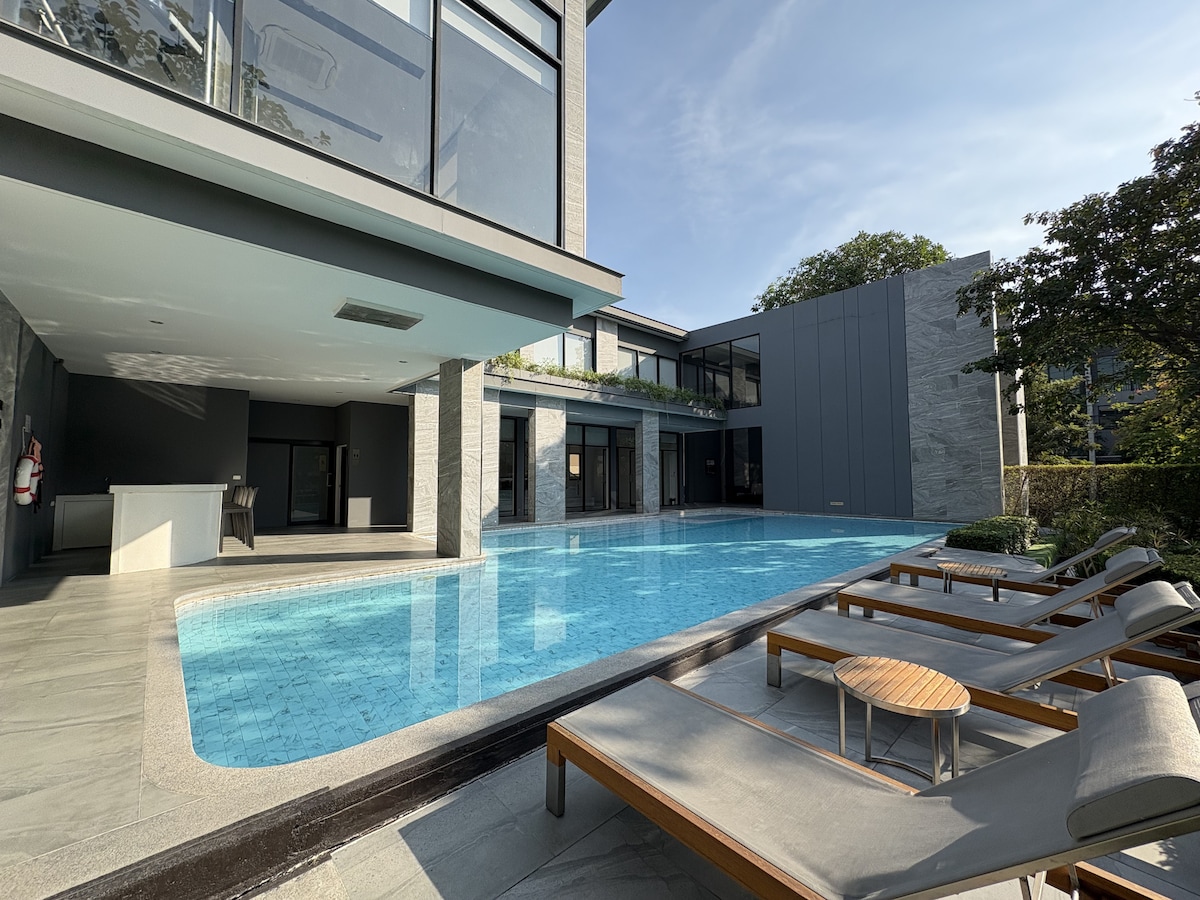 The outdoor area features a large swimming pool with clear blue water, surrounded by comfortable lounge chairs. The modern architecture of the adjoining building is highlighted by expansive glass walls, allowing views of the serene pool and landscaped surroundings.